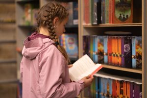 A school girl holding a book in her hands, standing near a bookshelf filled with books.